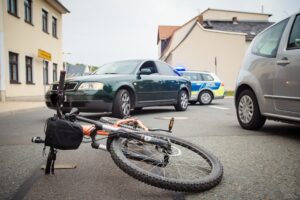 Unfallsituation auf der Straße, ein Fahrrad liegt im Vordergrund und im Hintergrund ein kaputtes Auto und ein Auto der Polizei